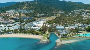 An aerial view of a coastal resort with multiple buildings, a beach, and a clear blue water body. - OCEAN VIEWS 10A pool & Spa Wi-Fi (Airlie Beach)