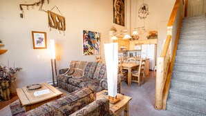 A cozy living room with patterned sofas, a wooden coffee table, and a dining area with a kitchen in the background. - Family-Friendly Canmore Crossing Condo | Mountain Views | Sleeps 7 (Canmore)