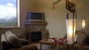 A cozy living room with a stone fireplace, a television on a wooden stand, and a view of mountains through a large window. - Family-Friendly Canmore Crossing Condo | Mountain Views | Sleeps 7 (Canmore)