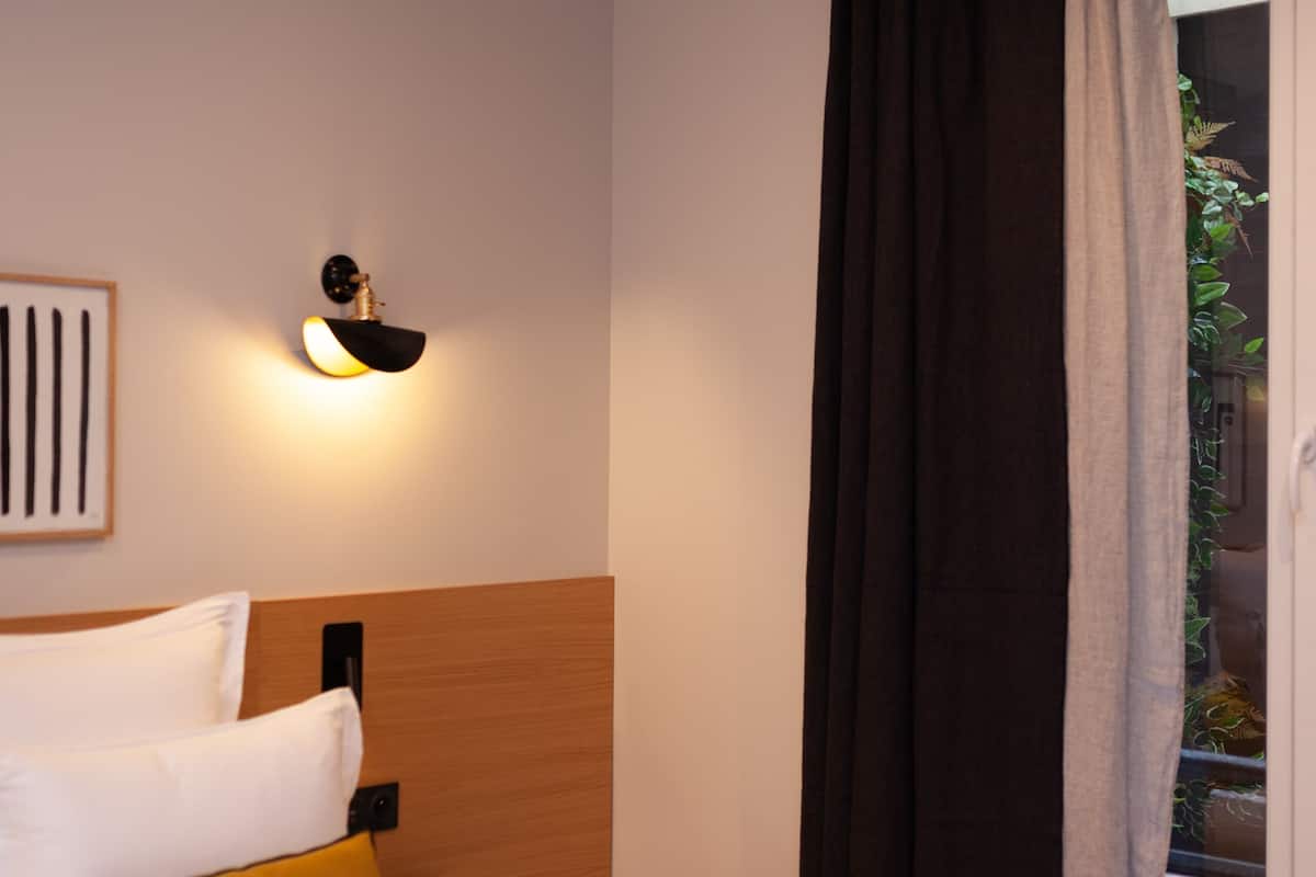 A hotel room with a bed, a nightstand, a wall-mounted light, and a window with curtains.