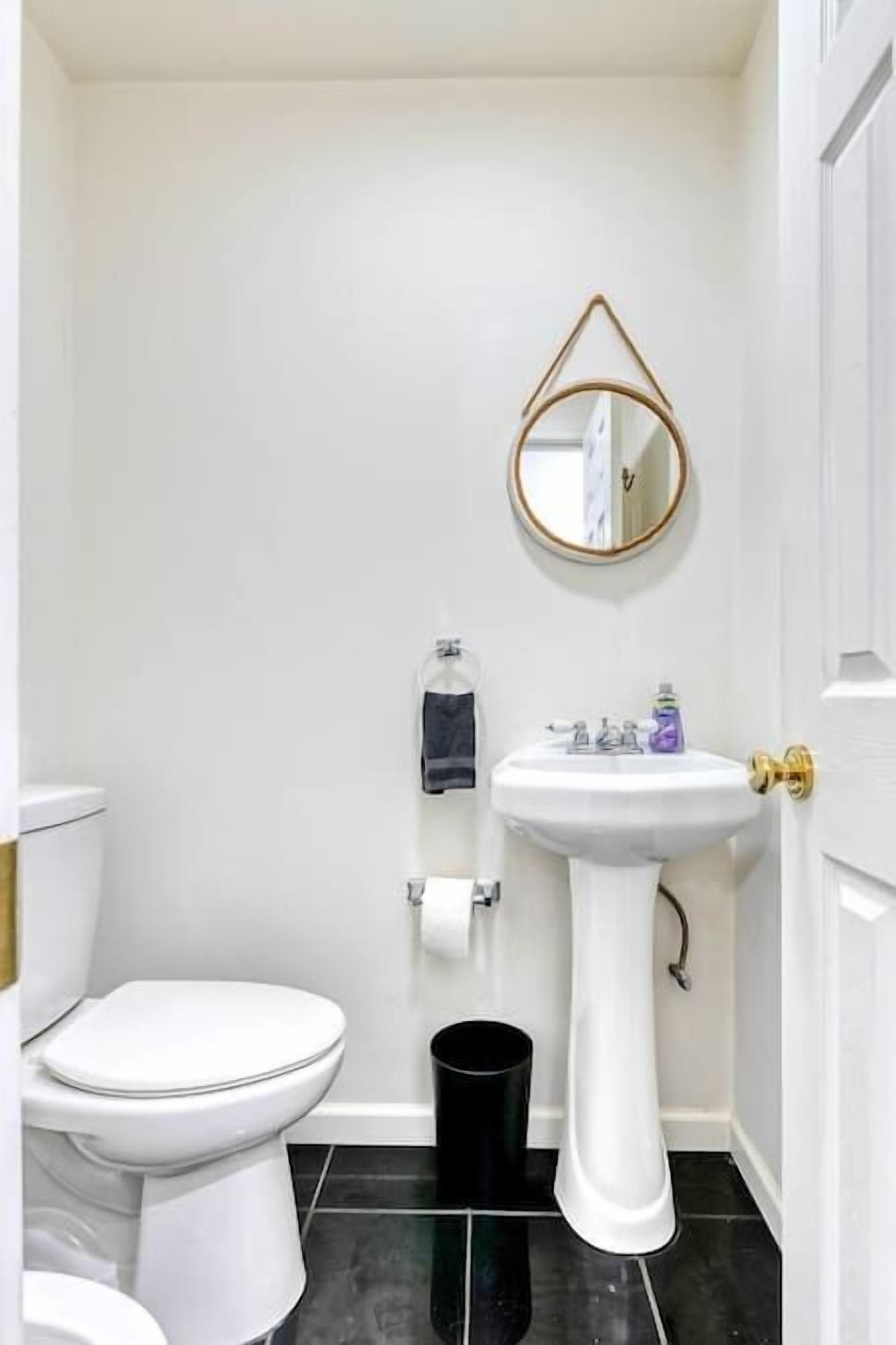 Une petite salle de bain blanche, avec des toilettes blanches, un lavabo sur colonne, une poubelle noire et un miroir encadré d’or accroché au mur.
