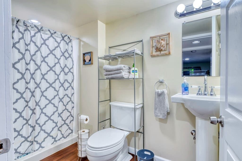 A bathroom with a shower curtain, toilet, shelf with towels, sink, and mirror.