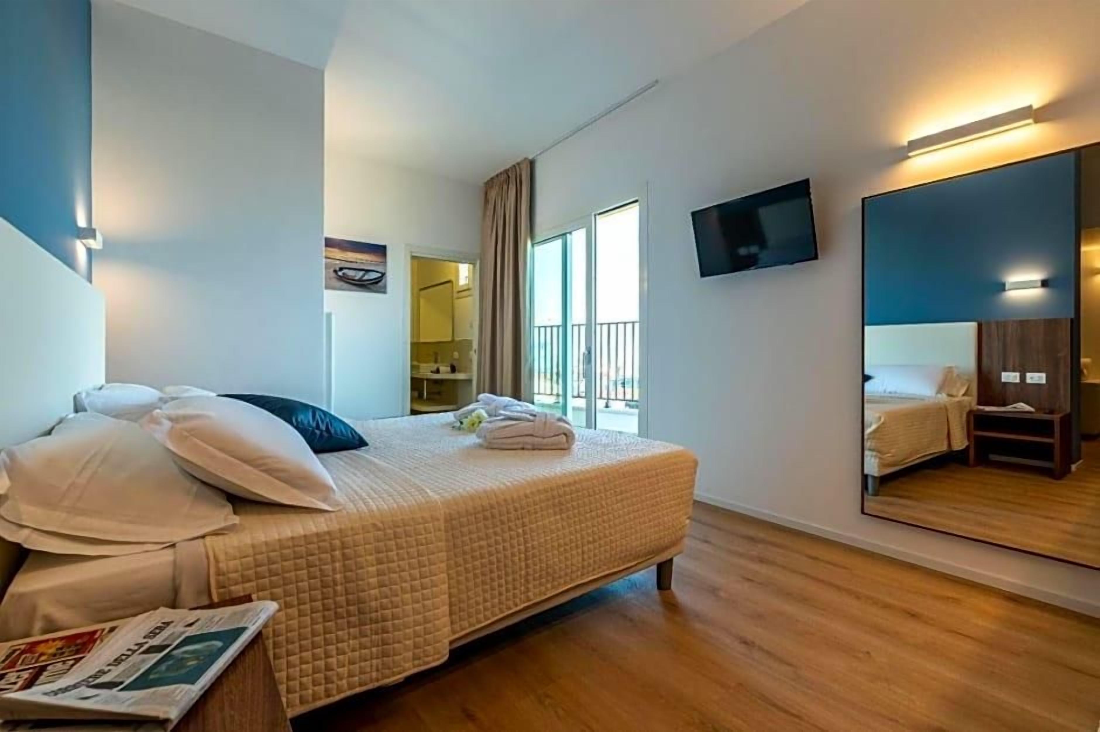 A hotel room with a bed, a flat-screen TV, a large mirror, and a window with curtains.