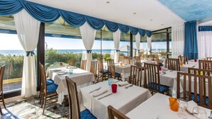 Un restaurant avec des tables dressées pour le service, de grandes fenêtres ornées de rideaux bleus et une vue sur la mer.