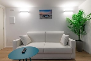 A white sofa with a remote control on a blue table, a potted plant, and a sunset painting on the wall. - Hotel Mondial (Jesolo)