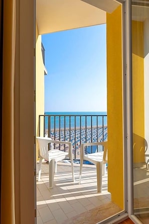 Ein Balkon mit Blick auf das Meer und den Strand, weiße Plastikstühle und eine Glastür.
