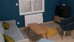 Un salón con una pared azul, un ventanal con marco blanco, un sofá gris, un sillón amarillo, un reposapiés azul, una mesa de centro de madera y un televisor sobre un mueble de madera.