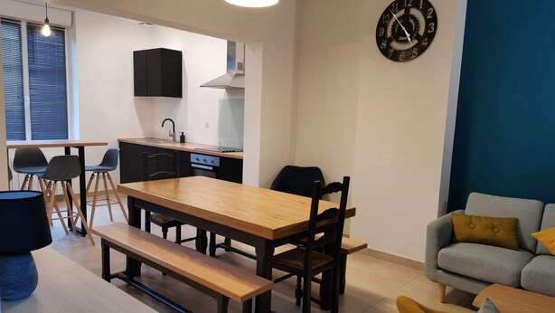 Una sala de estar moderna con una mesa de comedor de madera, sillas negras, un banco, un sofá gris con cojines amarillos y un reloj de pared.