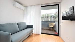 Ein modernes Wohnzimmer mit einem grauen Sofa, Holzboden, einer Schiebeglastür, die auf einen Balkon mit einem Sessel führt, und einem Wand-Fernseher.