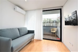 Een moderne woonkamer met een grijze bank, een houten vloer, een schuifdeur van glas naar een balkon met een stoel en een hangende televisie.