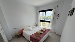 A bedroom with a bed, a small bedside table, a window with curtains, and a view of greenery outside. - Modern apartment in superb residence with PISCINE-3 Min PLAGE ROSAS- Clim-Wifi (Roses)