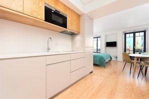 A modern kitchen with wooden cabinets, a built-in oven, and a sink. There is a dining area with a table and chairs, and a bed with a green bedspread.