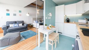 Ein kompakter Wohnbereich mit grauem Sofa, ottoman mit Streifenmuster, einem kleinen Esstisch mit Stühlen und einer Küchenzeile mit Spüle und Herd.