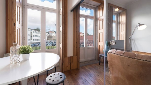 Une pièce avec une table ronde, une plante en pot, un canapé en cuir et de grandes fenêtres donnant sur des immeubles et un balcon.