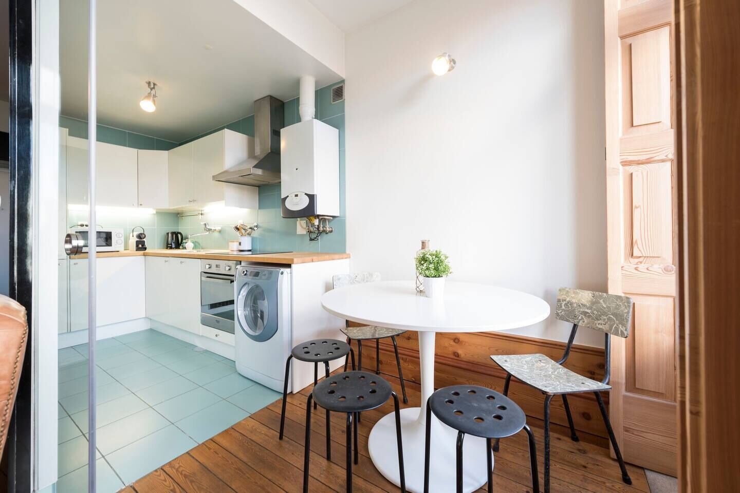 A compact kitchen with modern appliances, a dining area with a round table and four stools, and a wooden door.