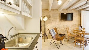 Una cocina con un lavaplatos de acero inoxidable, armarios blancos, una zona de comedor con sillas de madera y una pared de piedra.
