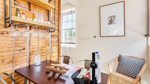 Een houten interieur met een eettafel, stoelen, een rieten fauteuil, een lijst met een afbeelding en planken met flessen en glazen.