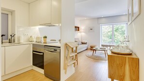 A modern kitchen with white cabinetry and a built-in dishwasher. There is a dining area with a wooden table and chairs, and a large window providing natural light. - Wood Premium Downtown Apartment by DA'HOME (Porto)