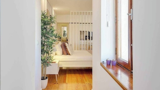 Ein schmaler Flur führt zu einem Schlafzimmer mit einem Bett, einer Zimmerpflanze und einem Fenster mit Jalousien.