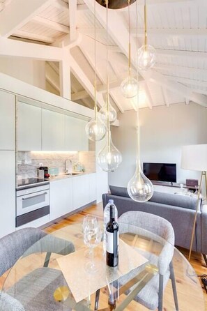 Une cuisine moderne avec des meubles blancs, un coin repas doté d’une table et de chaises en verre, et une grande fenêtre offrant une vue sur le ciel.