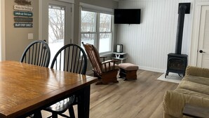 A cozy living room with a wooden dining table, chairs, a fireplace, and a television mounted on the wall. - Cozy & Private Lakeside Cabin on Lake Irene – Swim, Fish, Relax (Parkers Prairie Douglas County)