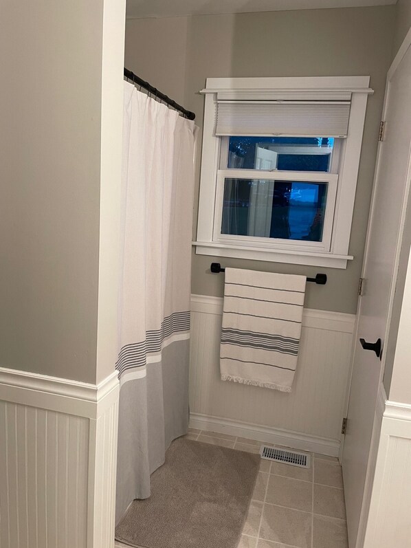Un baño pequeño con cortina de ducha, ventana, toallero y una alfombra pequeña.