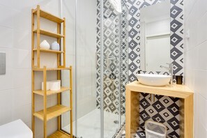 A modern bathroom with a glass-enclosed shower, a wooden shelf with toiletries, a sink with a mirror, and patterned tiles.