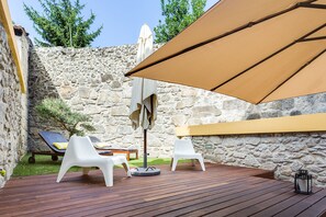 A wooden deck with white chairs and a table, a stone wall, and a large beige umbrella.