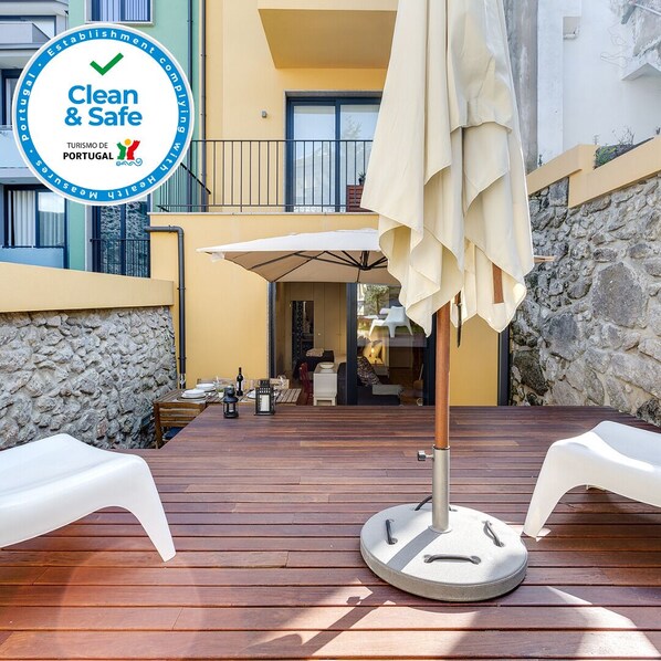 A wooden deck with a round table and a white umbrella, overlooking a building with a stone wall and a balcony.