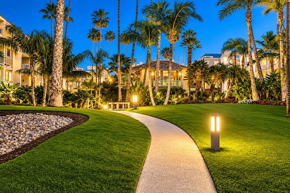A well-lit pathway leading to a building with multiple floors, surrounded by palm trees and a landscaped garden.