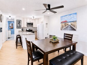 A modern kitchen with white cabinets, a microwave, and a refrigerator. There's a dining area with a wooden table and chairs, and a ceiling fan above. - 2BR Condo at North Coast Village | B116 (Oceanside)