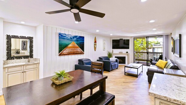 A spacious living room with a dining area, a ceiling fan, a large painting, and a TV. - 2BR Condo at North Coast Village | B116 (Oceanside)