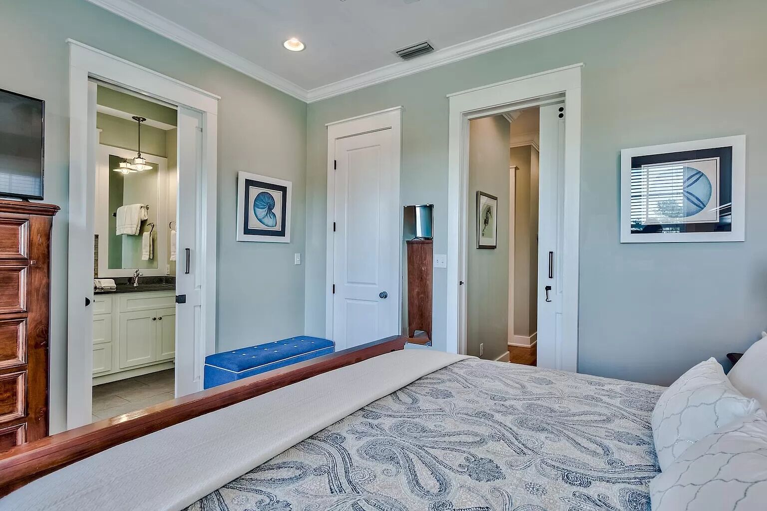 Une chambre à coucher comprenant un lit, un banc, une armoire et une salle de bain visible à travers une porte ouverte.