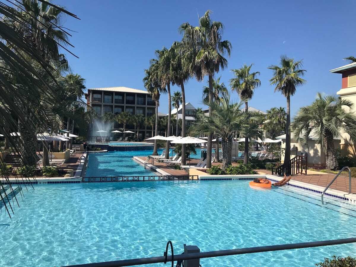 A swimming pool with lounge chairs and umbrellas, surrounded by palm trees and a building in the background.