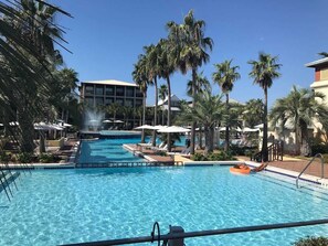Una piscina con sillas de descanso y sombrillas, rodeada de palmeras y un edificio al fondo.
