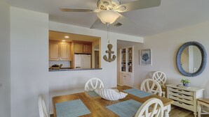 Un coin repas avec une table en bois, des chaises blanches et des sets de table bleus. On y trouve un ventilateur de plafond, une cuisine visible à travers une porte ouverte et un miroir sur le mur.