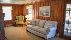 Un salón acogedor con un sofá color beige, un sillón estampado y una mesita de noche con una lámpara. Un cuadro cuelga en la pared, y se ve una ventana con cortinas.
