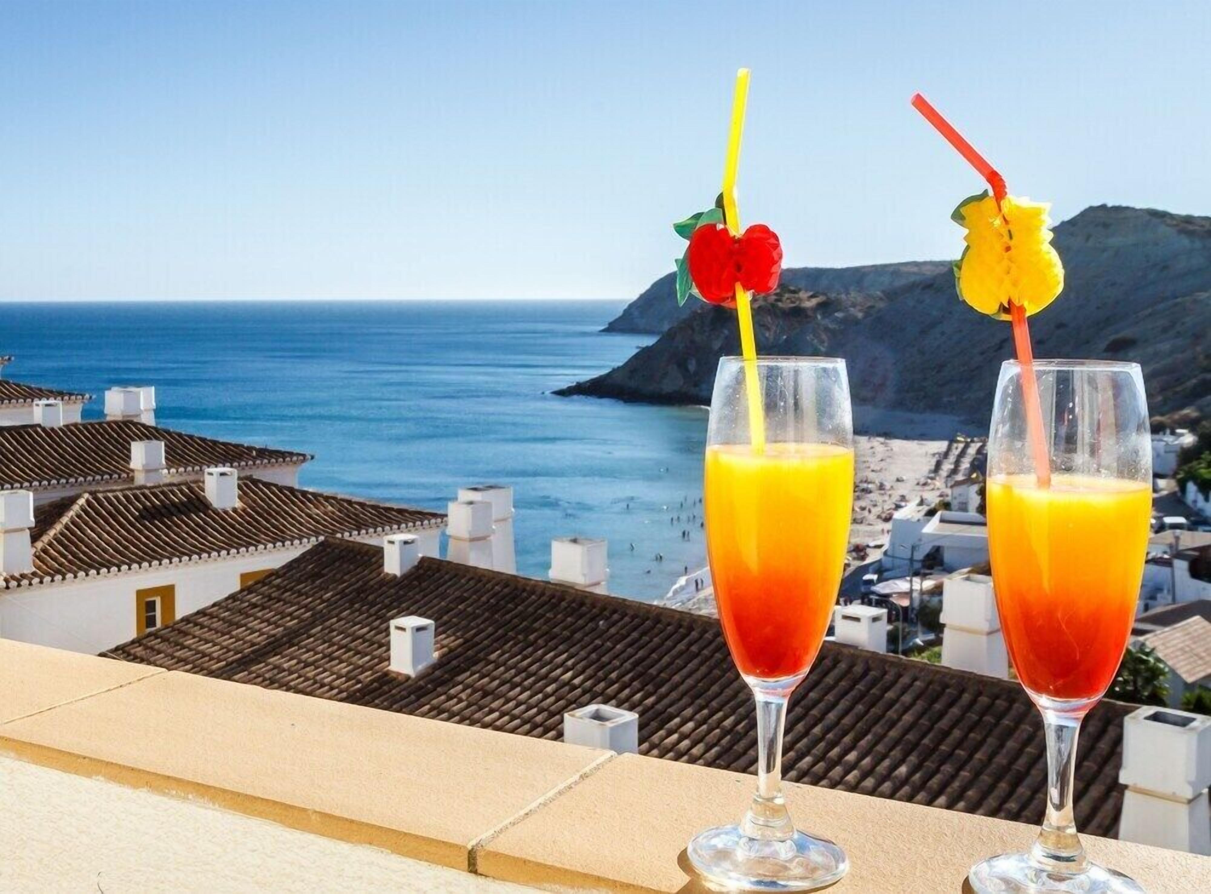 Deux verres de jus d’orange, décorés de morceaux de fruits, sont posés sur un balcon donnant sur une ville côtière, avec ses bâtiments et la mer.