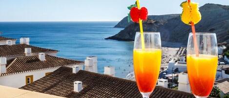 Deux verres de jus d’orange, décorés de morceaux de fruits, sont posés sur un balcon donnant sur une ville côtière, avec ses bâtiments et la mer.