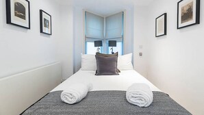 A neatly made bed with two pillows, a grey blanket, and two rolled towels. There are framed pictures on the walls and a window with curtains. - Lovely flat in Chelsea (London)