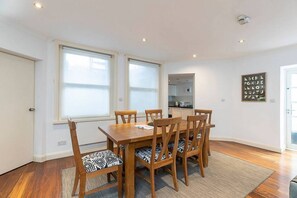 A dining area with a wooden table and chairs, a kitchen in the background, and a large window with blinds.