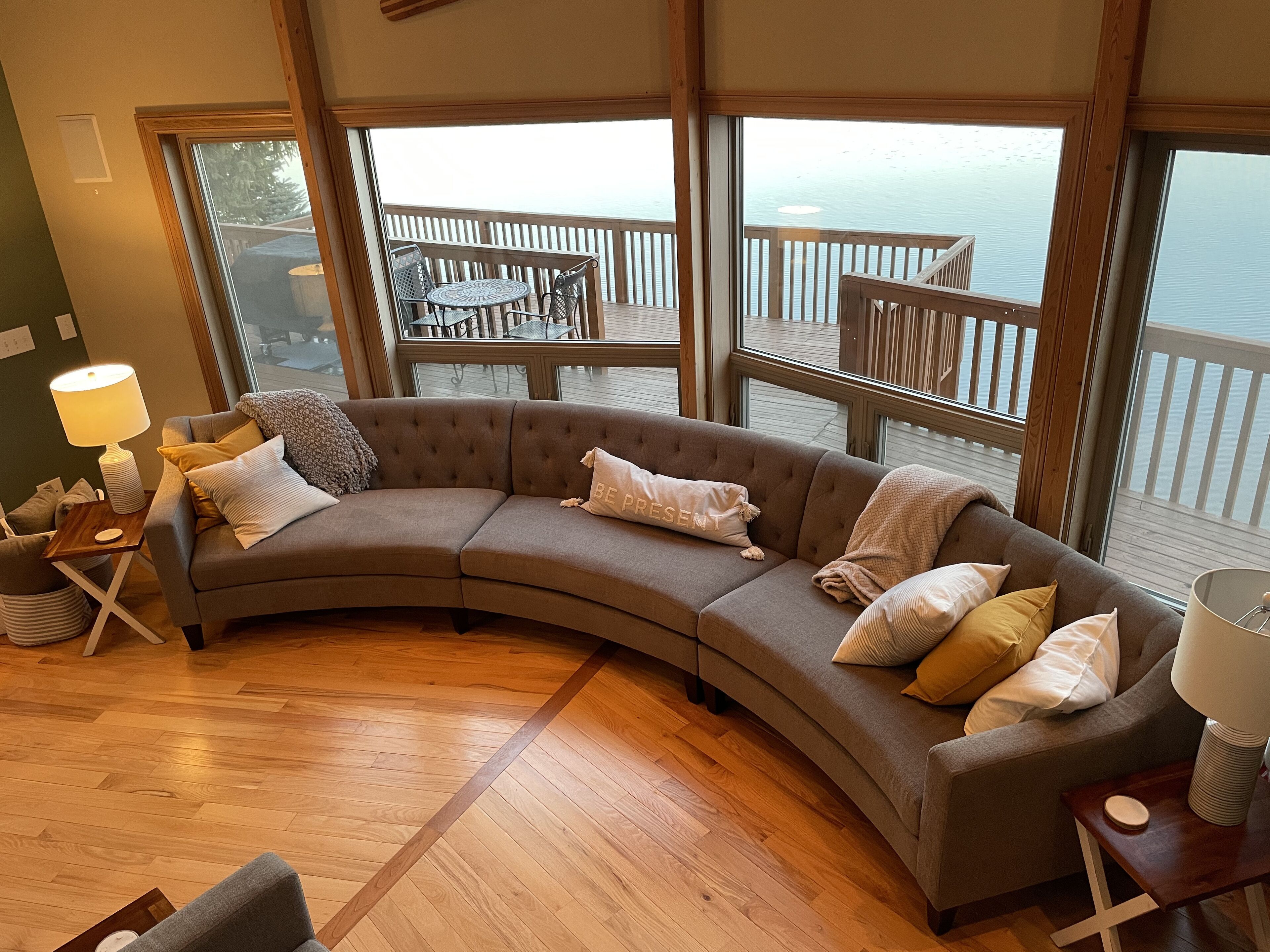 A curved sofa with multiple pillows, a small table with a lamp, and a view of a wooden deck and water through large windows.