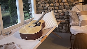Ein Zimmer mit Steinwand, einer Gitarre auf einem Tisch, einem Stuhl und Blick durch große Fenster.