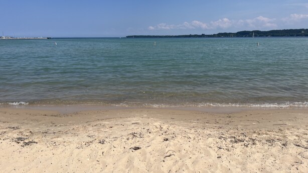 Ein Strand mit klarem Wasser, sandigem Ufer und einer entfernten Küstenlinie mit Gebäuden und Bäumen.