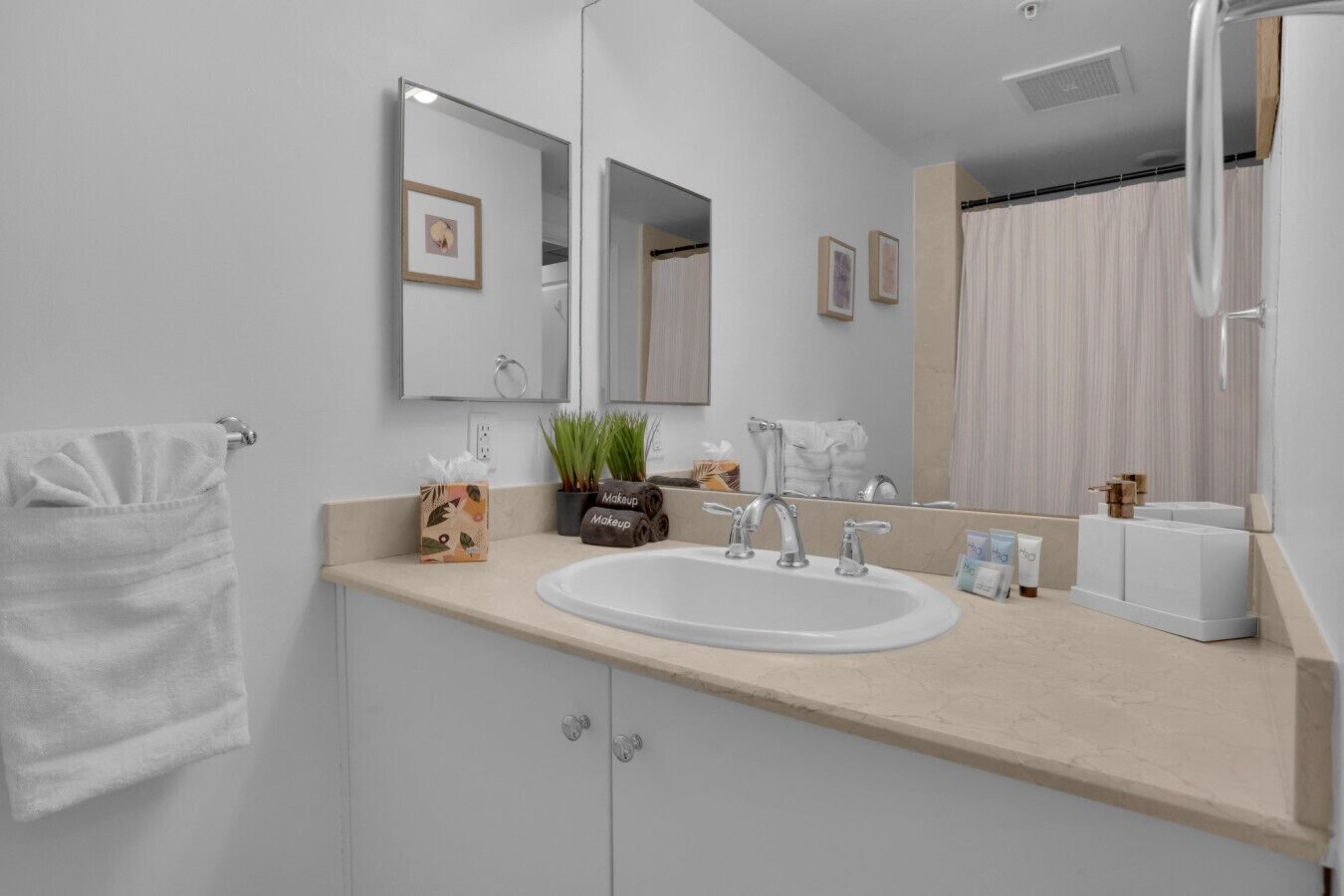 Een moderne badkamer met een wit wastafel, twee spiegels, een handdoekhouder, een make-up set en een potplant.