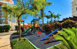 Un parque infantil con toboganes y estructuras para escalar, rodeado de árboles y un edificio al fondo.