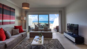 A modern living room with a sofa, a coffee table, and a TV. There is a large window with a view of the ocean. - Praia del Rey 3 Bedroom Villa (Obidos)