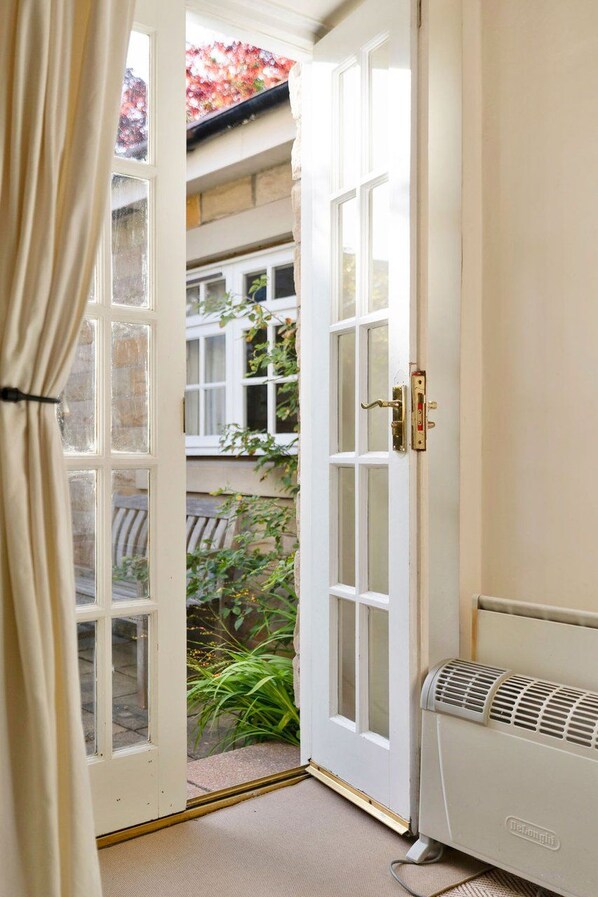 A room with a white door, a radiator, and a view of a garden with plants and a building in the background.