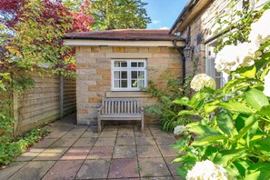 A stone building with a bench and a window, surrounded by plants and a wooden fence.