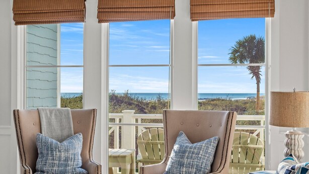 Una habitación con dos sillones color beige, una mesa de madera y vistas a la playa y al océano a través de ventanas grandes con persianas marrones.